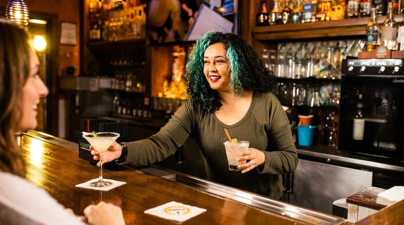 A smiling bartender with teal-streaked hair serves a margarita to a customer at a lively bar.