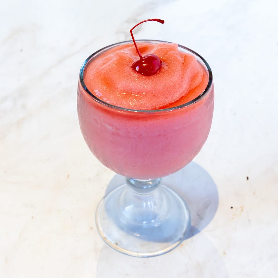 Pink and red frozen drink in a stemmed glass, topped with a maraschino cherry.