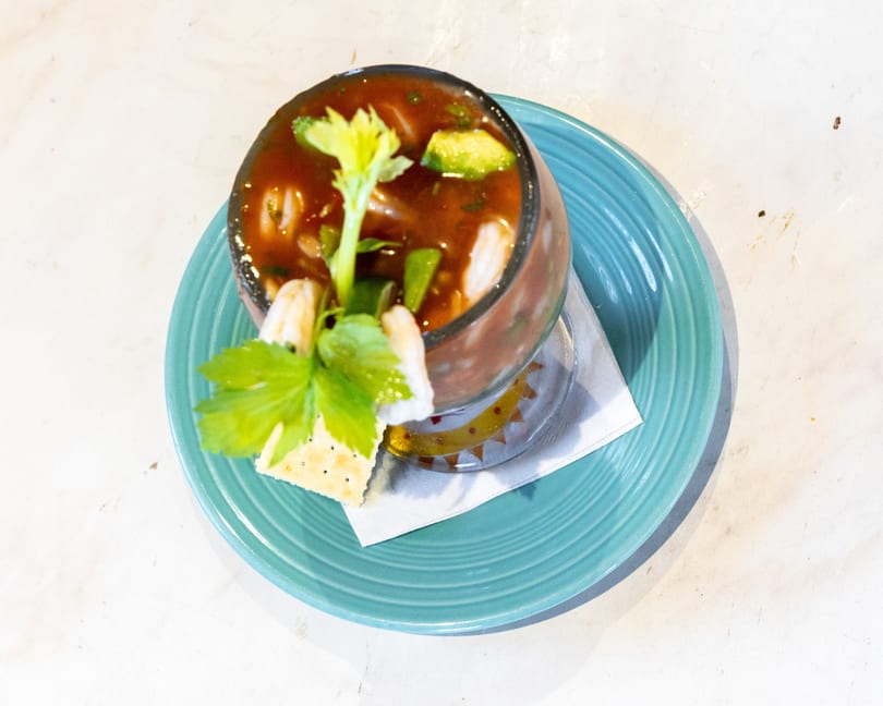 A vibrant shrimp cocktail with avocado and celery in a glass, served on a blue plate with crackers.