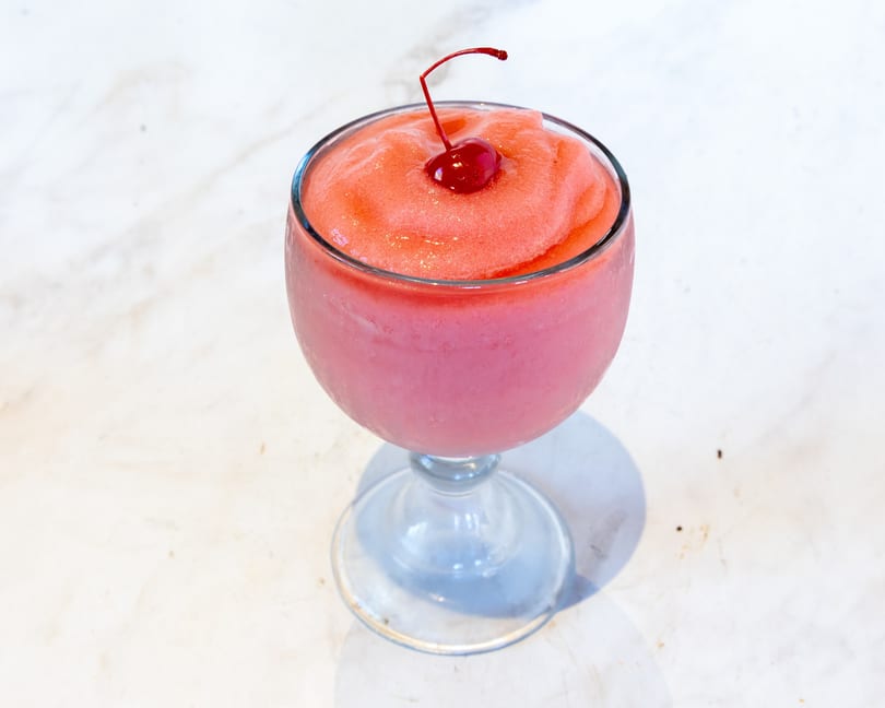 Frozen pink and red drink in a glass, garnished with a maraschino cherry.
