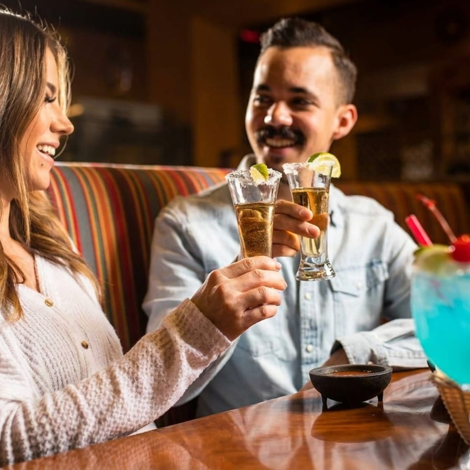 Smiling couple toasting with tequila shots at a vibrant restaurant table with colorful drinks.