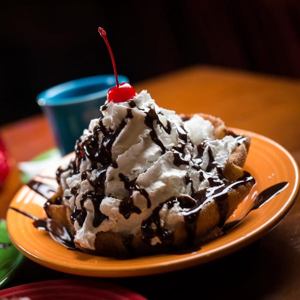 Golden fried dessert topped with whipped cream, chocolate drizzle, and a bright red cherry.