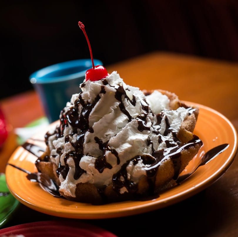 Golden fried dessert topped with whipped cream, chocolate drizzle, and a bright red cherry.