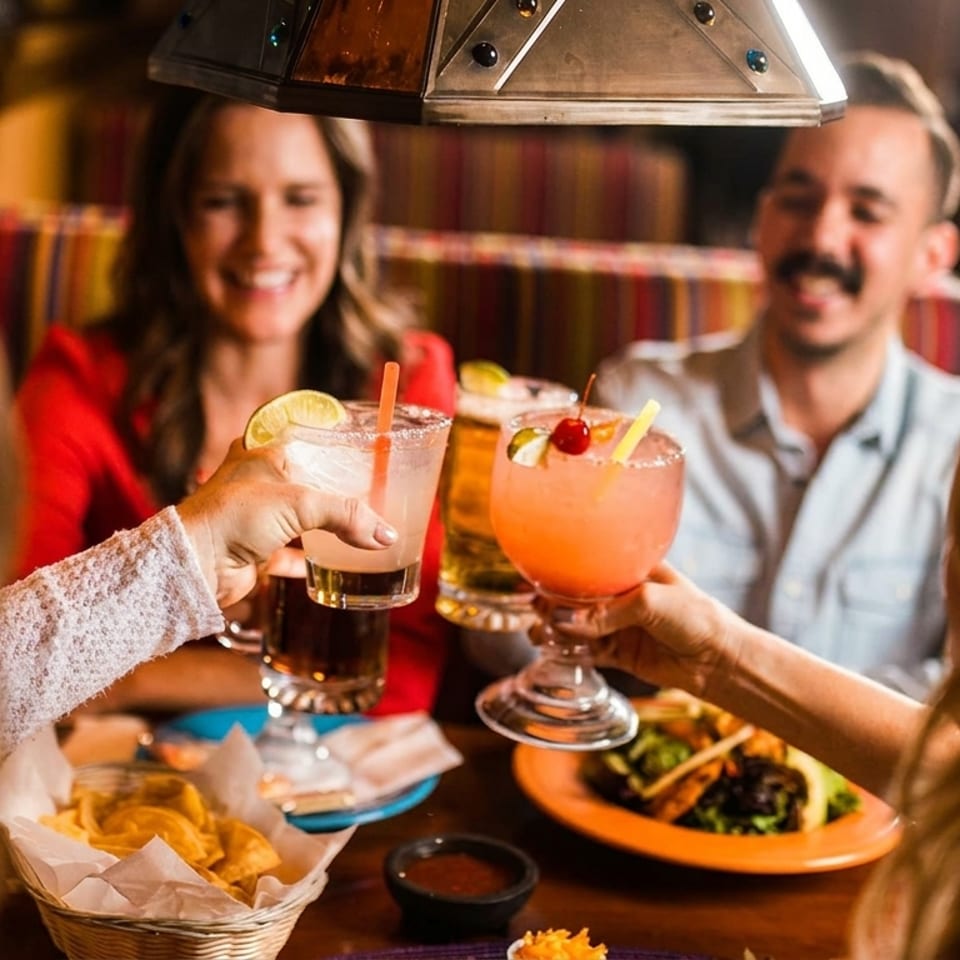 Four people clinking colorful cocktails and drinks at a restaurant table with food.