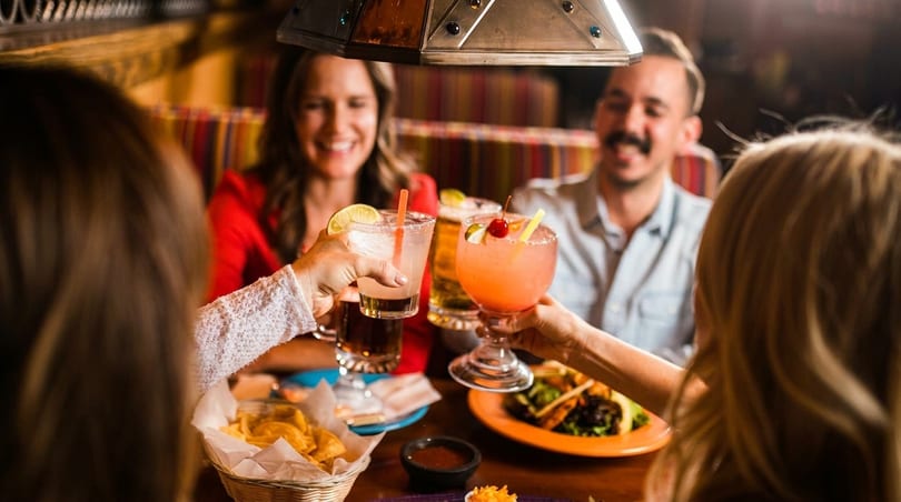 Four people clinking colorful cocktails and drinks at a restaurant table with food.