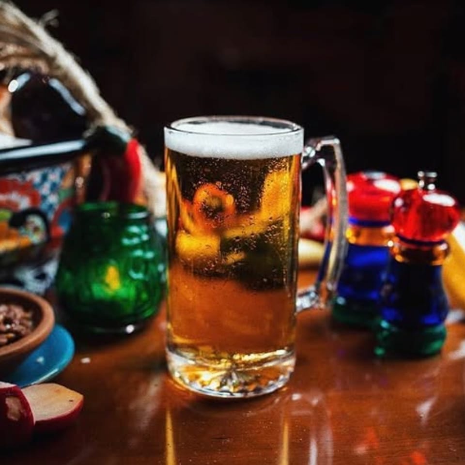 Mug of foamy beer surrounded by colorful Mexican dishes, chips, and condiments on a wooden table.