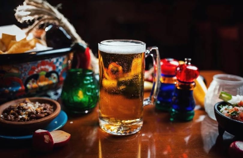 Mug of foamy beer surrounded by colorful Mexican dishes, chips, and condiments on a wooden table.