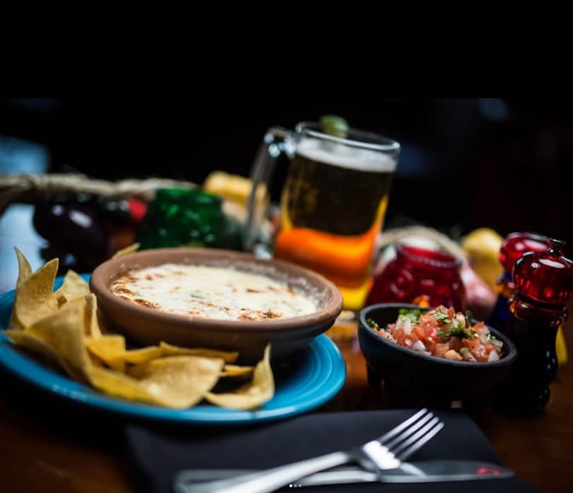 Queso with tortilla chips, pico de gallo, and a beer on a restaurant table.